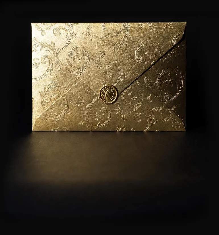 A luxurious gold envelope with subtle embossed floral patterns, sealed with a pointed flap, centred against a deep black background.
